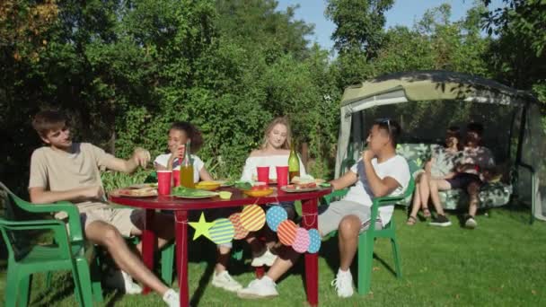 Συντροφιά αστείων φίλων που κάθονται στο τραπέζι και απολαμβάνουν υπαίθρια καλοκαιρινό πικνίκ γεύμα — Αρχείο Βίντεο