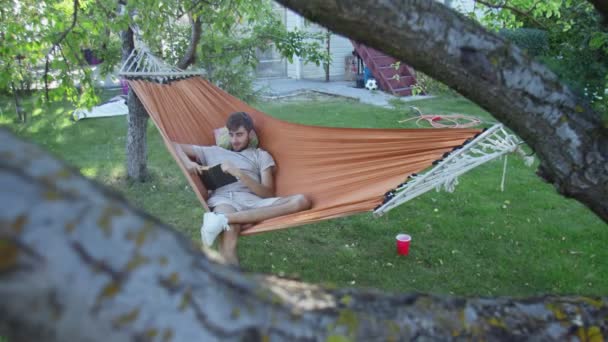 Młody człowiek czytający książkę leżąc w hamaku na podwórku — Wideo stockowe