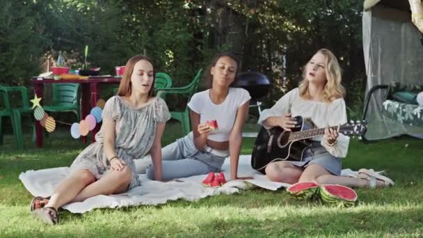 Νεαρή γυναίκα παίζει κιθάρα και δύο γυναίκες τραγουδούν και τρώνε καρπούζι. Οι φίλες κάθονται σε μια κουβέρτα στο πάρκο. — Αρχείο Βίντεο