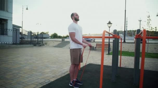 Молодий м'язистий чоловік виконує вправи з еластичною смугою на відкритому повітрі. Тренування — стокове відео