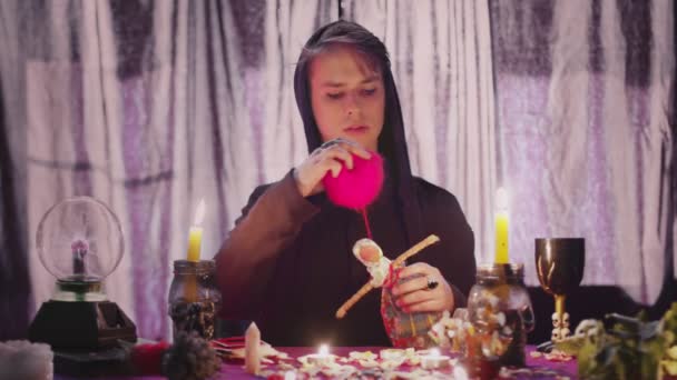 Czarna czarownica voodoo wykonująca rytualne i rzucające zaklęcie przekłuwające lalkę z igłami siedzącymi w ciemnym pokoju z płonącymi świecami. — Wideo stockowe