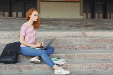 Dalgın öğrenci dizüstü kullanarak merdivenlerde oturan