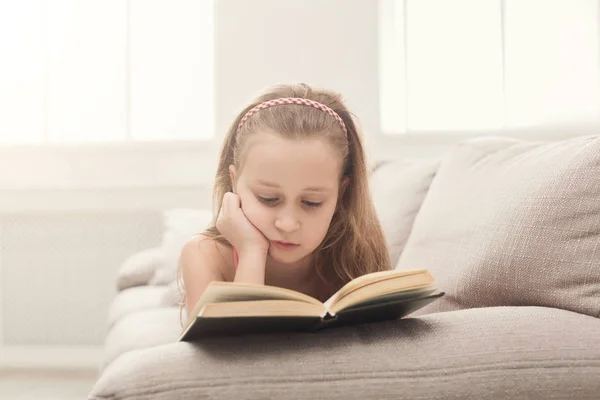 Menina bonita ler livro, deitado no sofá em casa — Fotografia de Stock