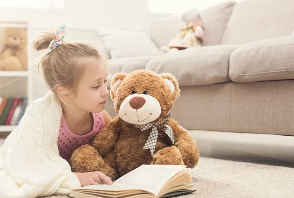 Happy little girl and her teddy bear reading book