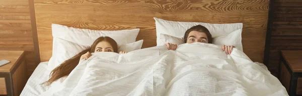 Zszokowany mężczyzna i kobieta ukrywa się pod kocem w ich własnym łóżku, w domu — Zdjęcie stockowe