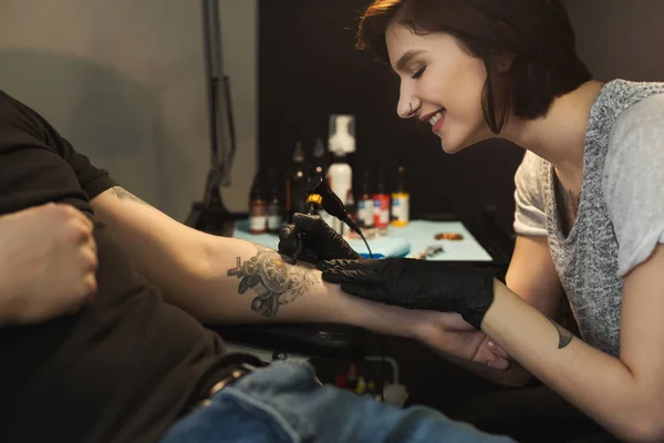 Young woman tattooist doing tattoo on male arm