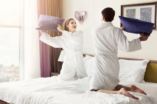 Joven pareja feliz en albornoz almohadas de lucha en la cama — Foto de Stock