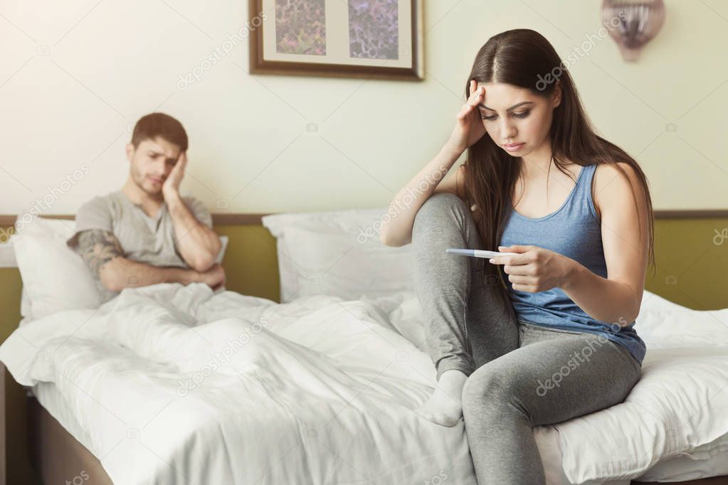 Upset woman sitting on bed and looking at pregnancy test