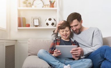 Father and son using tablet together on sofa at home clipart