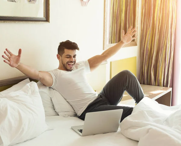 Mladý muž pomocí přenosného počítače, zatímco sedí na posteli — Stock fotografie