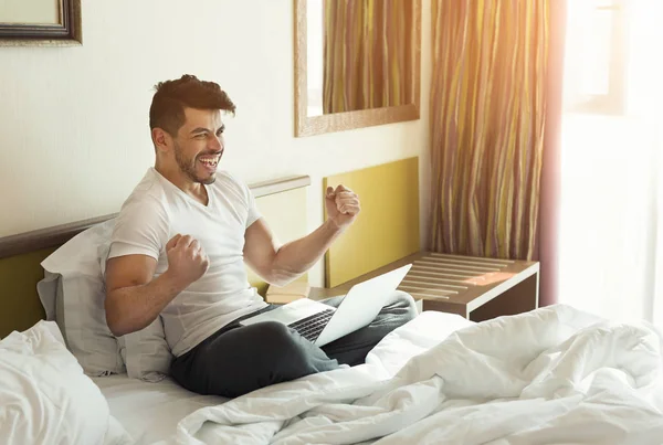 Hombre joven usando el ordenador portátil mientras está sentado en una cama —  Fotos de Stock