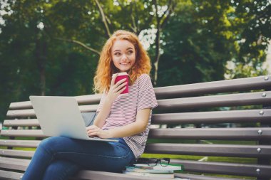 Kızıl saçlı öğrenci kız gülümseyerek parkta kahve içer