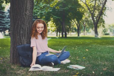 Kızıl saçlı kız açık havada kitap okuma ve bir ağacın altında oturup dizüstü bilgisayar kullanarak