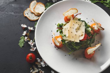 Siyah arka plan üzerine beyaz plaka restoran yemek çiçekler, yeşillik ve domates ile dekore edilmiştir. Ispanak, roka, peynir, somon ve baget, üstten görünüm ile salata