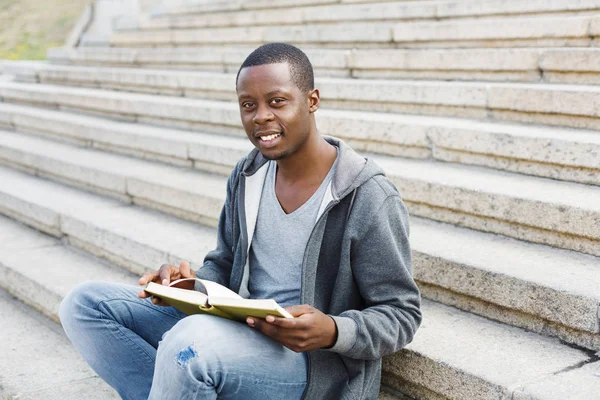 Sonriente estudiante afroamericano leyendo libro en escaleras universitarias — Foto de Stock