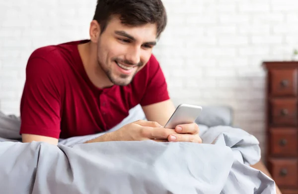 Bonito homem usando smartphone enquanto deitado na cama — Fotografia de Stock