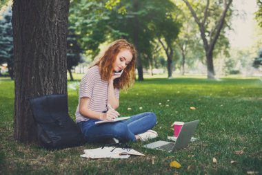 Bir ağacın altında oturan çalışan genç redhead öğrenci kız