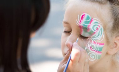 Little girl getting her face painted by face painting artist. clipart