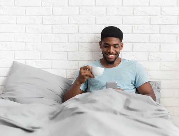 Μαύρος χρησιμοποιώντας smartphone και να πίνει καφέ στο κρεβάτι — Φωτογραφία Αρχείου