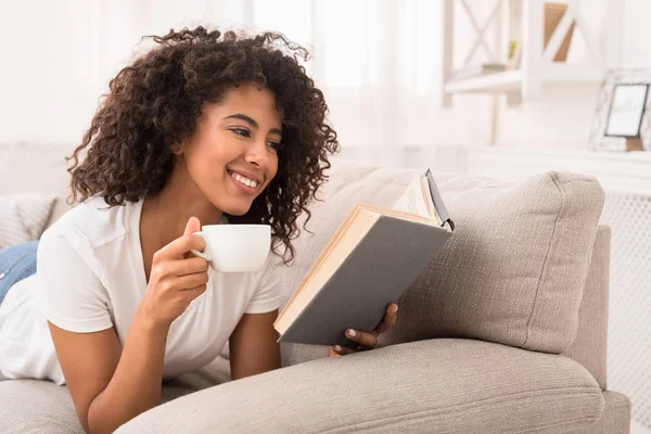 Mujer feliz bebiendo té y leyendo libro en el sofá — Foto de Stock