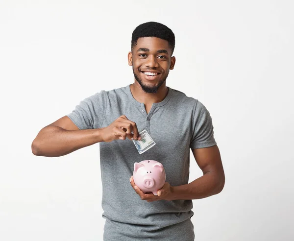 Sorridente uomo africano-americano mettere soldi nel suo salvadanaio — Foto Stock