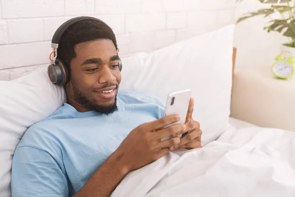 Schwarzer Mann mit Kopfhörer schaut Smartphone im Bett — Stockfoto