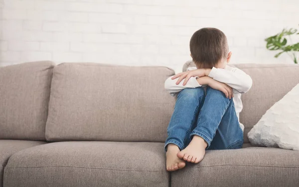 Offended boy hugging his knees in tear, sitting on sofa, copy space