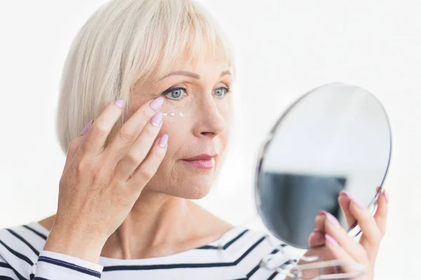 Belle femme âgée tenant miroir et appliquant de la crème visage — Photo