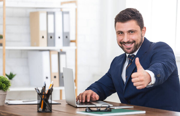 Businessman showing thumb up, using laptop in office