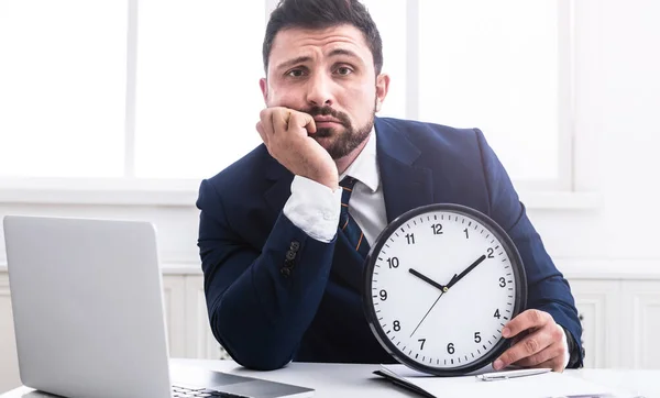 Tired businessman holding clock, sitting at workplace — Stock Photo, Image