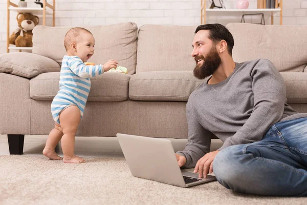 Junger Vater arbeitet am Laptop und kümmert sich um seinen kleinen Sohn — Stockfoto