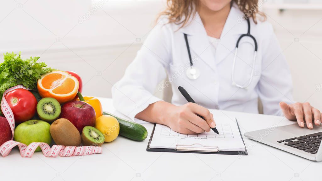 Nutritionist working in office. Doctor writing diet plan on table and using laptop, panorama, copy space
