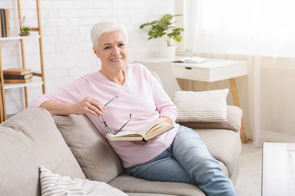 Старшая женщина отдыхает на диване и читает книгу — стоковое фото