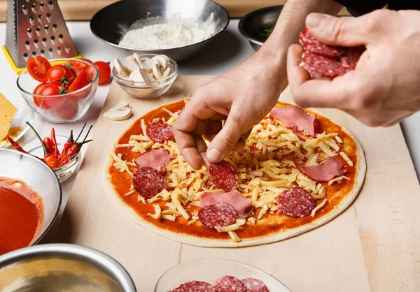 Mann bereitet Pizza zu, dekoriert sie mit Salami und Schinken — Stockfoto
