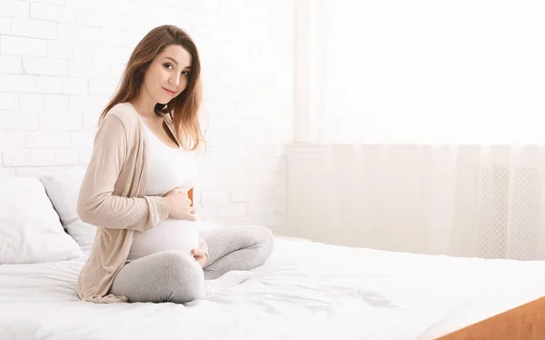 Mujer feliz embarazada tocando su vientre en la cama —  Fotos de Stock
