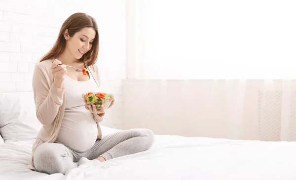 Feliz embarazada joven sentada y comiendo ensalada de frutas en el sofá en casa — Foto de Stock