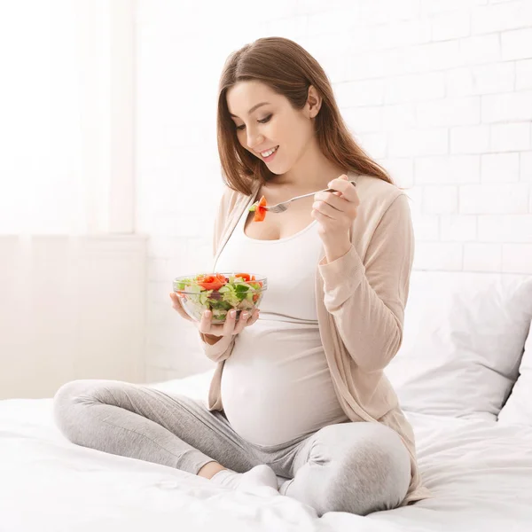 Feliz embarazada joven sentada y comiendo ensalada de frutas en el sofá en casa — Foto de Stock