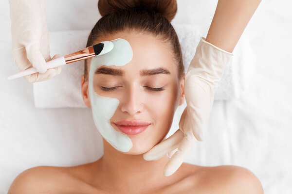 Beautician applying clay face mask on woman face