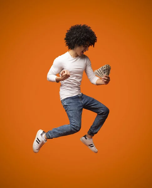 Возбужденный молодой человек прыгает с деньгами на оранжевом фоне — стоковое фото