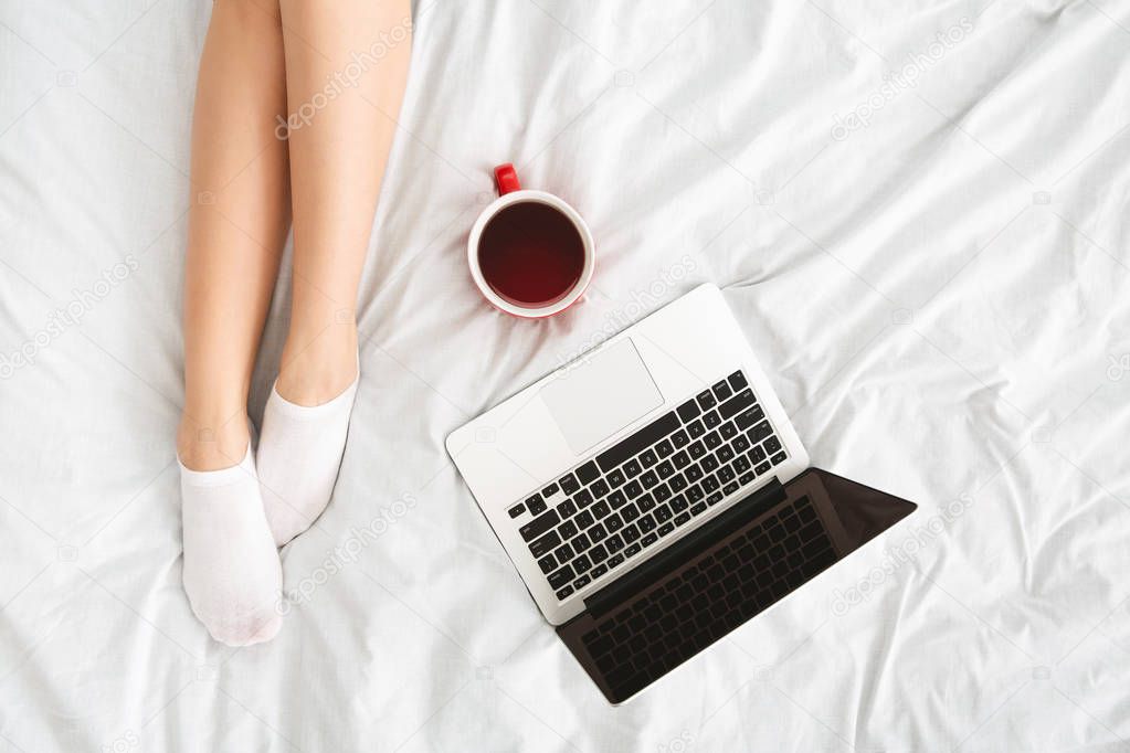 Top view on female legs, laptop and cup of coffee on bed