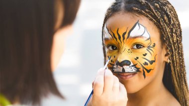 Adorable african-american girl getting tiger face painting clipart