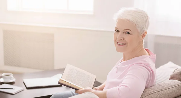 Портрет улыбающейся пожилой женщины с книгой на диване дома — стоковое фото