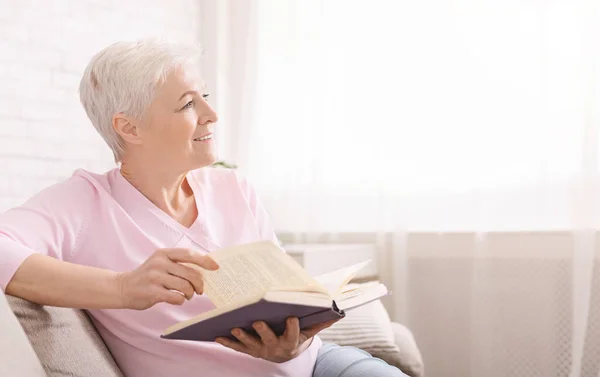 Sonhador mulher sênior relaxante em casa com livro favorito — Fotografia de Stock