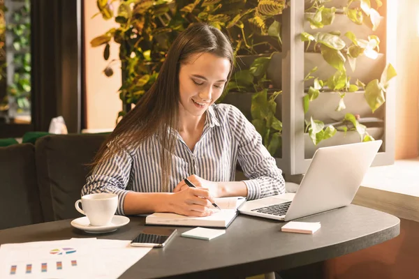 Young woman sitting at cafe in front of laptop and making notes — 스톡 사진