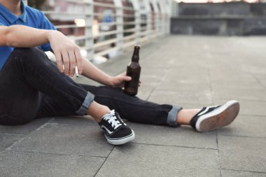 Genç adam sigara içiyor ve bira içiyor, yalnız oturuyor.