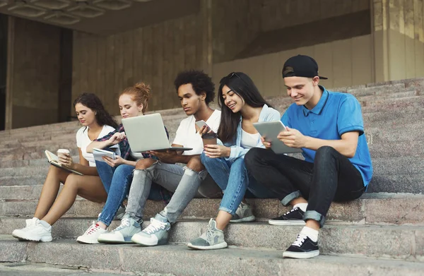 Grupo de diversos adolescentes que utilizan gadgets, sentado en escaleras al aire libre — Foto de Stock