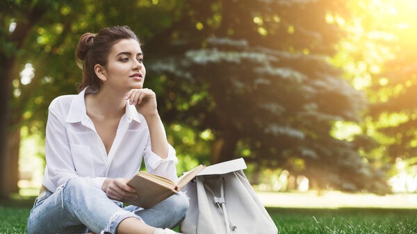 Outdoor photo of woman sitting on grass with book and dreaming