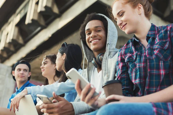 Amis adolescents multiethniques assis à l'extérieur, parlant et utilisant des gadgets — Photo