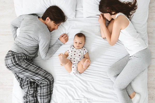 Tired parents sleeping on bed sides, baby playing in the middle