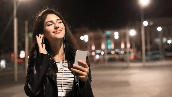 Laza lány élvezi a zene fülhallgató, séta az éjszakai városban — Stock Fotó